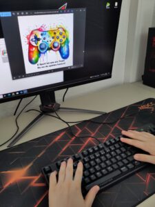 Teilnehmer*in hat Hände auf der PC Tastatur und designed ein Werbeschild für einen E-Sport-Wettbewerb. Auf dem Bildschirm ist ein bunter Controller zu sehen.